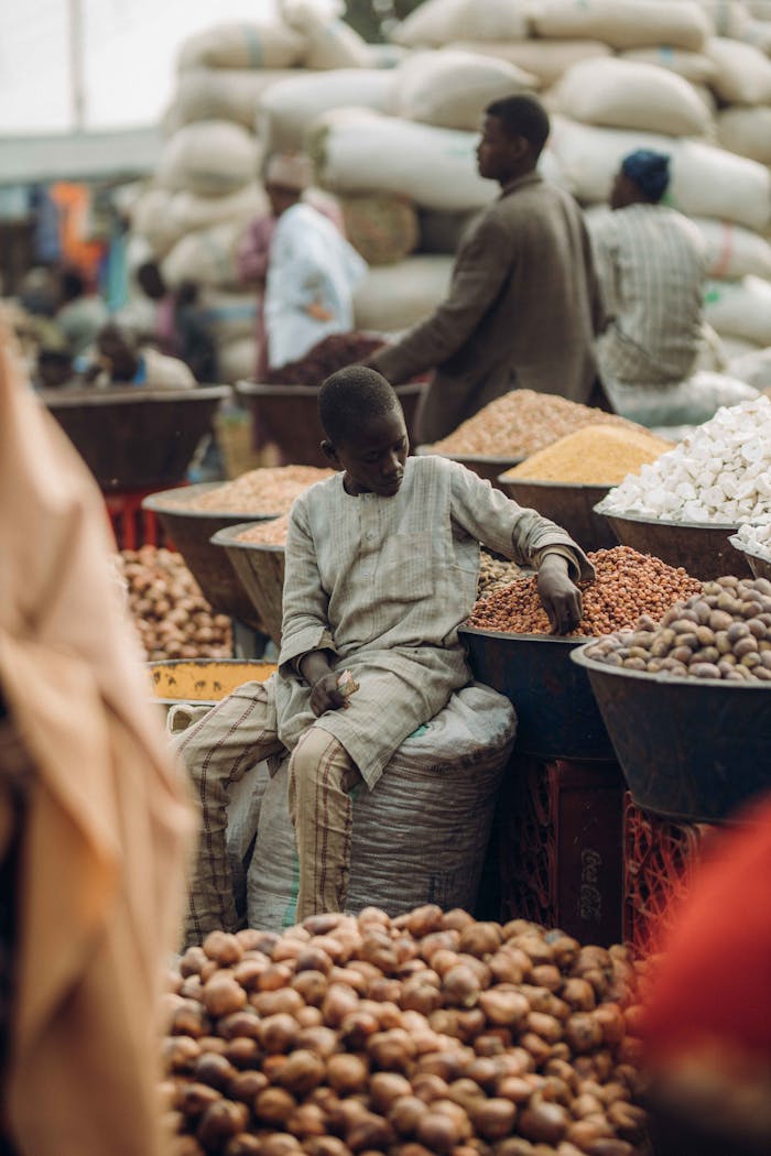 our-story-01 A young boy sits amidst a bustling market surrounded by sacks and bowls of grains and nuts.
