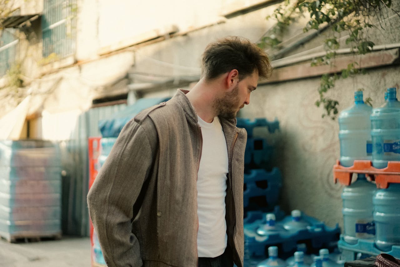 A young man in a jacket stands near stacked water bottles outdoors, urban setting.